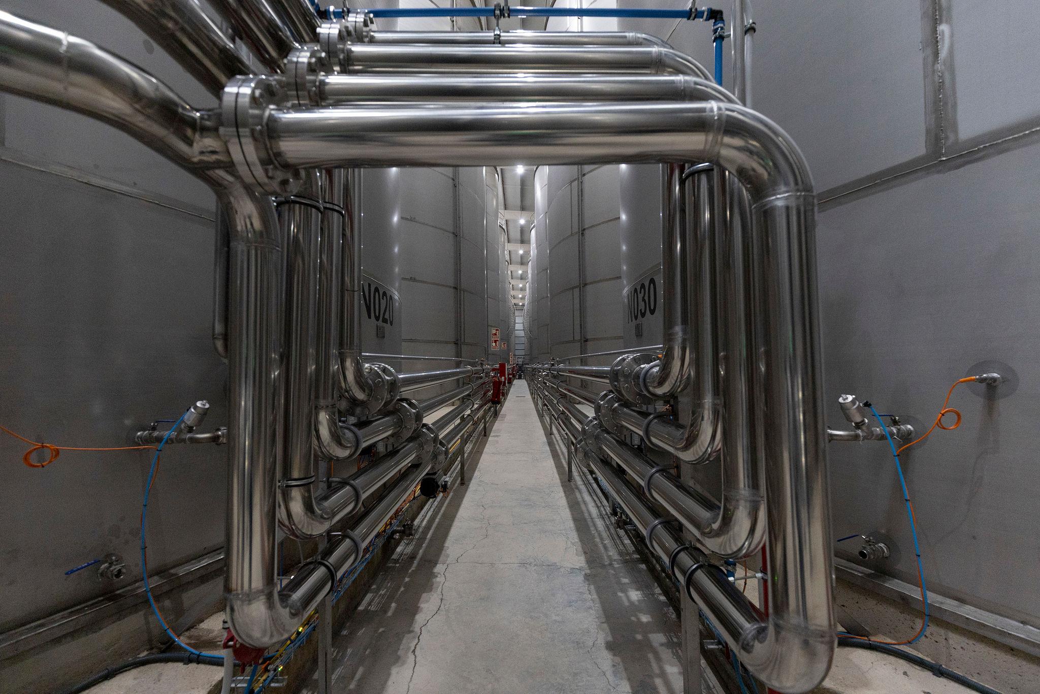 A corridor between two rows of numbered stainless steel storage tanks, with pipework arching overhead